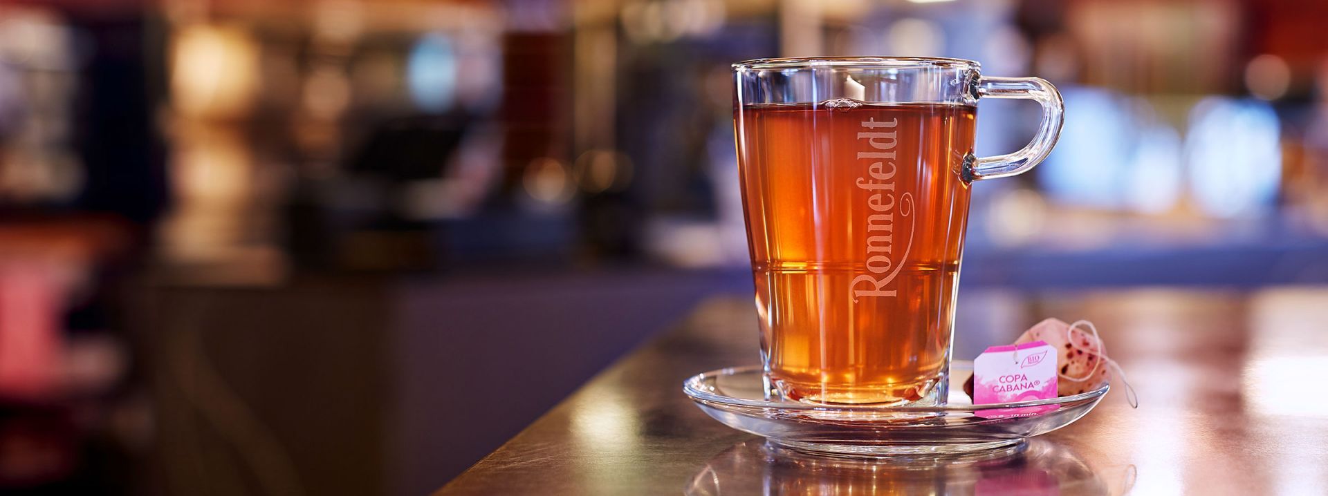 Glass cup of Ronnefeldt Copa Cabana tea on a reflective surface in a warmly lit setting.