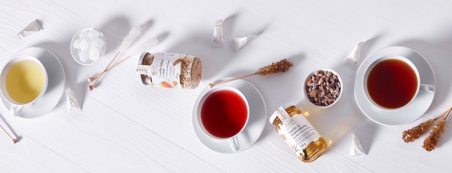 Three cups of tea in different colors with rock sugar sticks, sweeteners, and jars of flavored sugar and honey on a white wooden surface.