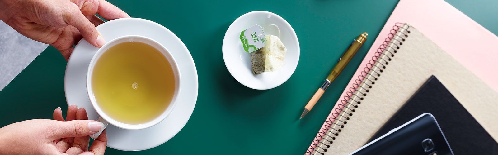 Cup of green tea on a green desk with a used tea bag, pen, notebooks, and phone.