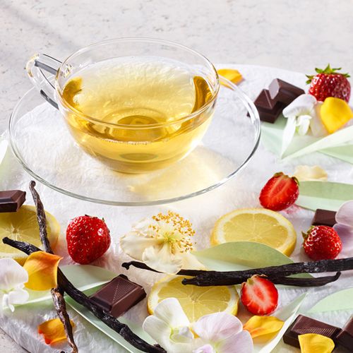 Glastasse mit grünem Tee auf einem Hintergrund mit Erdbeeren, Zitronenscheiben, Schokoladenstücken, Vanilleschoten, Blütenblättern und grünen Teeblättern.