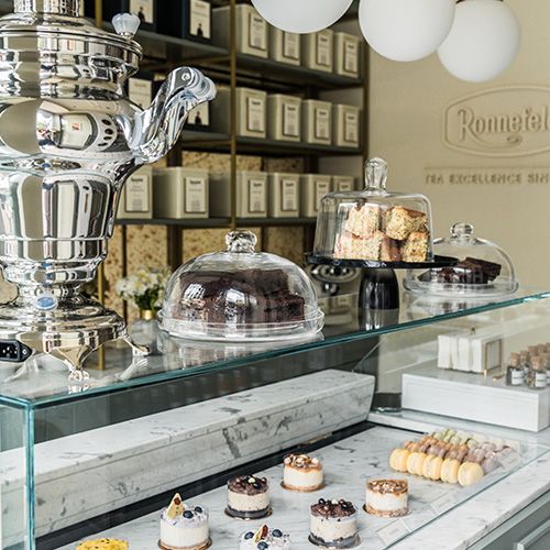 Teegeschäft mit einer Auswahl an feinen Desserts und Gebäck in einer Glasvitrine, einem glänzenden Samowar und Regalen mit Ronnefeldt Teedosen im Hintergrund