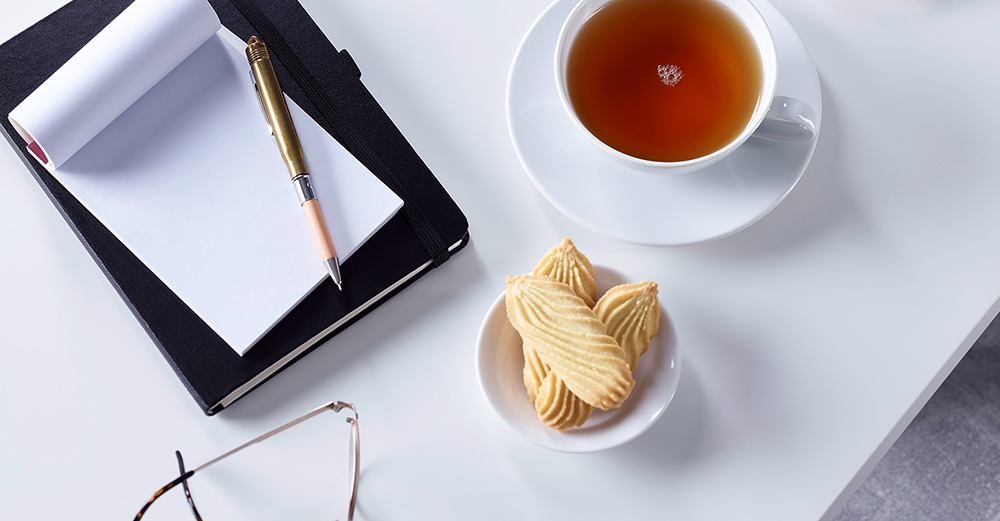 Tasse schwarzer Tee mit Buttergebäck, daneben ein Notizbuch mit Stift und eine Brille auf weißem Tisch.