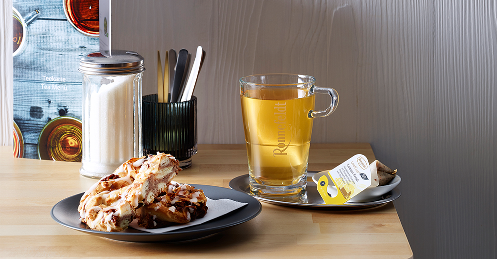 Tasse mit dem Tee, Teebeutel „Lemon Fresh“, Gebäck auf einem Teller, Zuckerbehälter, Besteck und Teekarte auf einem Holztisch.