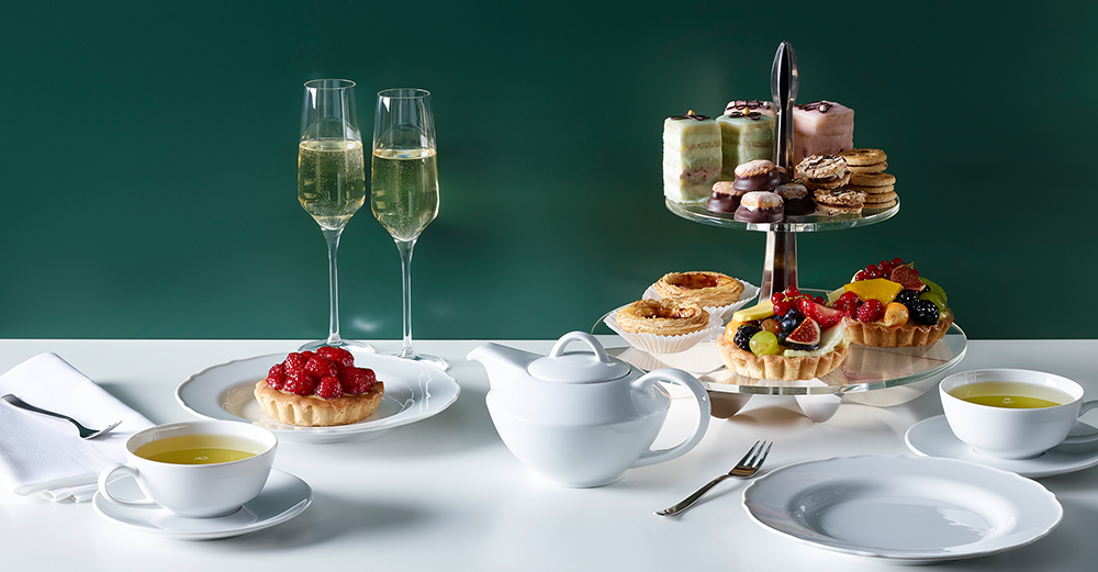 Elegant gedeckter Tisch mit weißem Porzellan, zwei Gläsern Sekt, einer Etagere voller Kuchen und Gebäck sowie zwei Tassen grünem Tee.