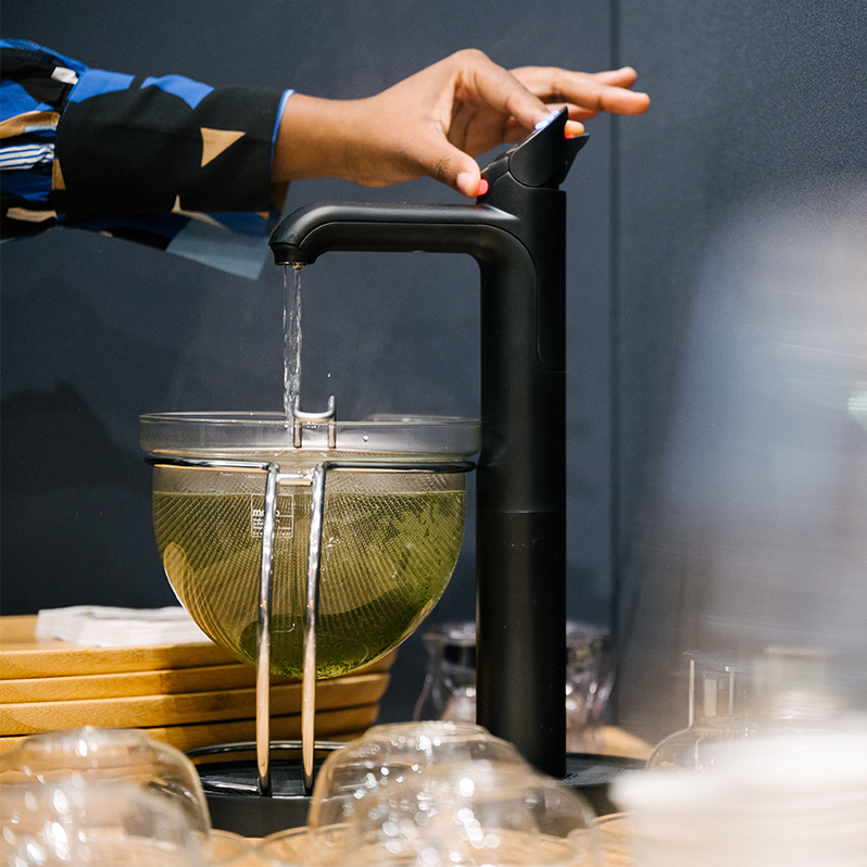 Eine Person füllt heißes Wasser aus einem modernen schwarzen Wasserspender in eine Glaskanne mit Teesieb, in dem grüner Tee zieht.