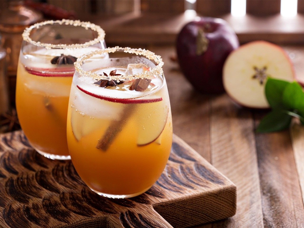 Zwei Tee-Cocktails mit Eiswürfeln, Apfelscheiben, Zimtstangen und Sternanis auf einem Holzbrett, im Hintergrund frische Äpfel