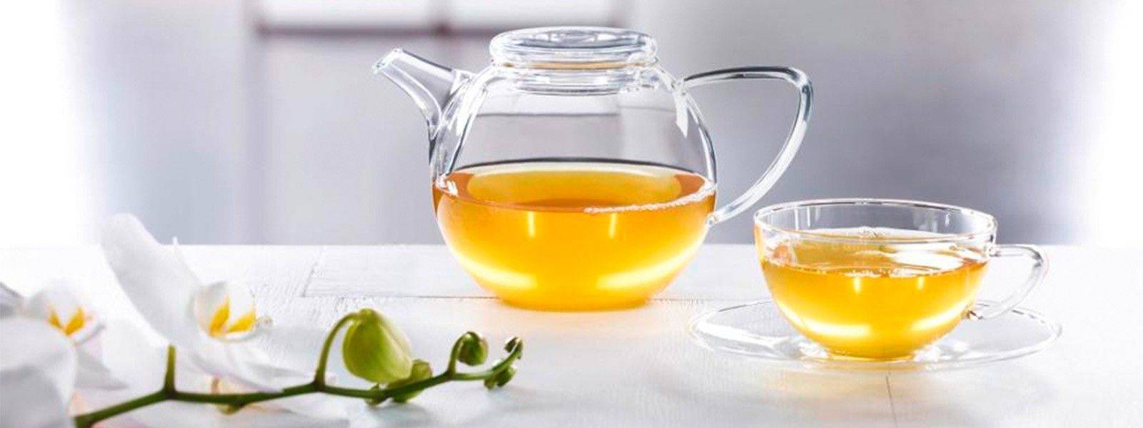 Glasteekanne und Teeglas mit grünem Tee auf weißem Tisch mit Orchideenblüten