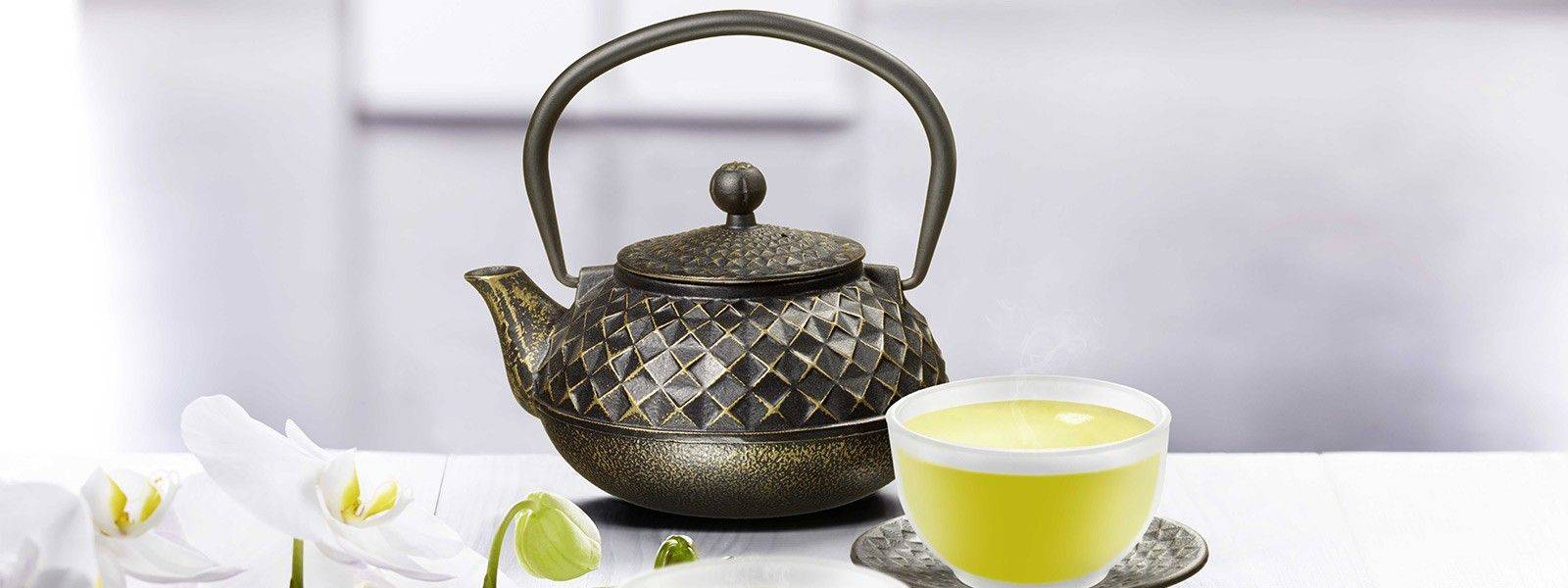 Gusseiserne Teekanne mit filigranem Muster neben dampfender Tasse grünem Tee und weißen Orchideen