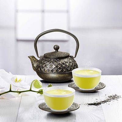 Zwei Tassen grüner Tee mit gusseiserner Teekanne und Orchideenblüte auf weißem Tisch