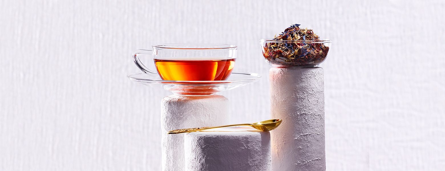 Glas-Tasse mit dem Tee auf einer weißen Säule, daneben eine Glasschale mit bunter Teemischung und ein goldener Teelöffel im Vordergrund