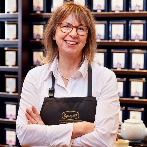 Lächelnde Mitarbeiterin im Ronnefeldt Brandshop mit schwarzer Schürze vor einem Regal mit Teedosen