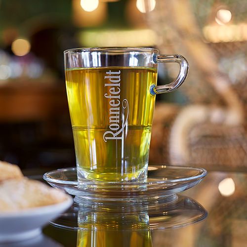 Ronnefeldt Glastasse mit Kräutertee auf einer Glas-Untertasse, im Hintergrund ein gemütliches Café mit warmem Licht.