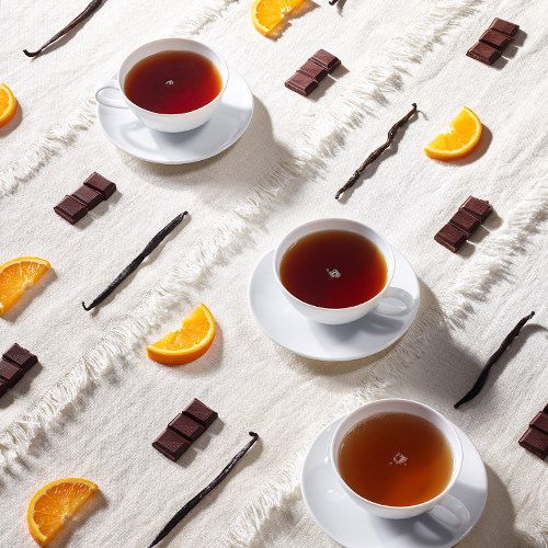 Drei weiße Teetassen mit schwarzem Tee auf heller Tischdecke, dekoriert mit Orangenspalten, Schokoladenstücken und Vanilleschoten.