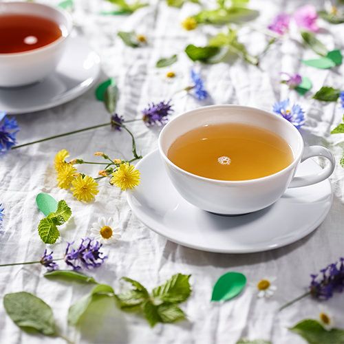 Weiße Teetasse mit Kräutertee auf einer weißen Tischdecke, dekoriert mit bunten Blüten, grünen Blättern und einer zweiten Teetasse im Hintergrund.