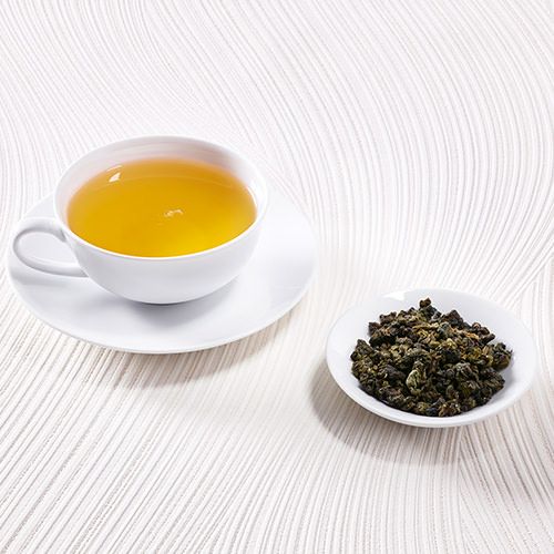 Weiße Teetasse mit goldgelbem Oolong-Tee auf einem hellen Hintergrund, daneben eine kleine Schale mit losem Oolong-Tee.