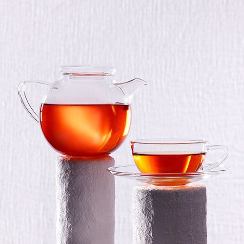 Glaskanne und Glastasse mit Rooibos-Tee auf weißen Säulen vor hellem Hintergrund.