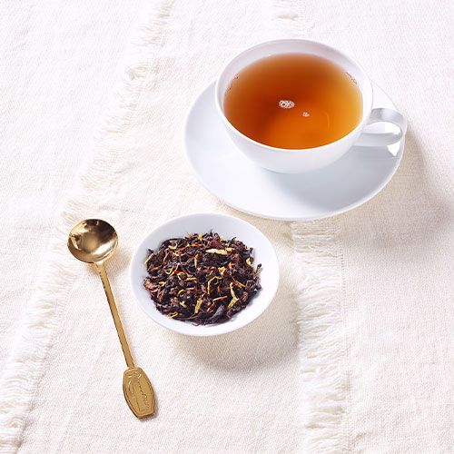 Weiße Teetasse mit schwarzem Tee auf heller Tischdecke, daneben eine kleine Schale mit losem Tee und ein goldener Teelöffel.